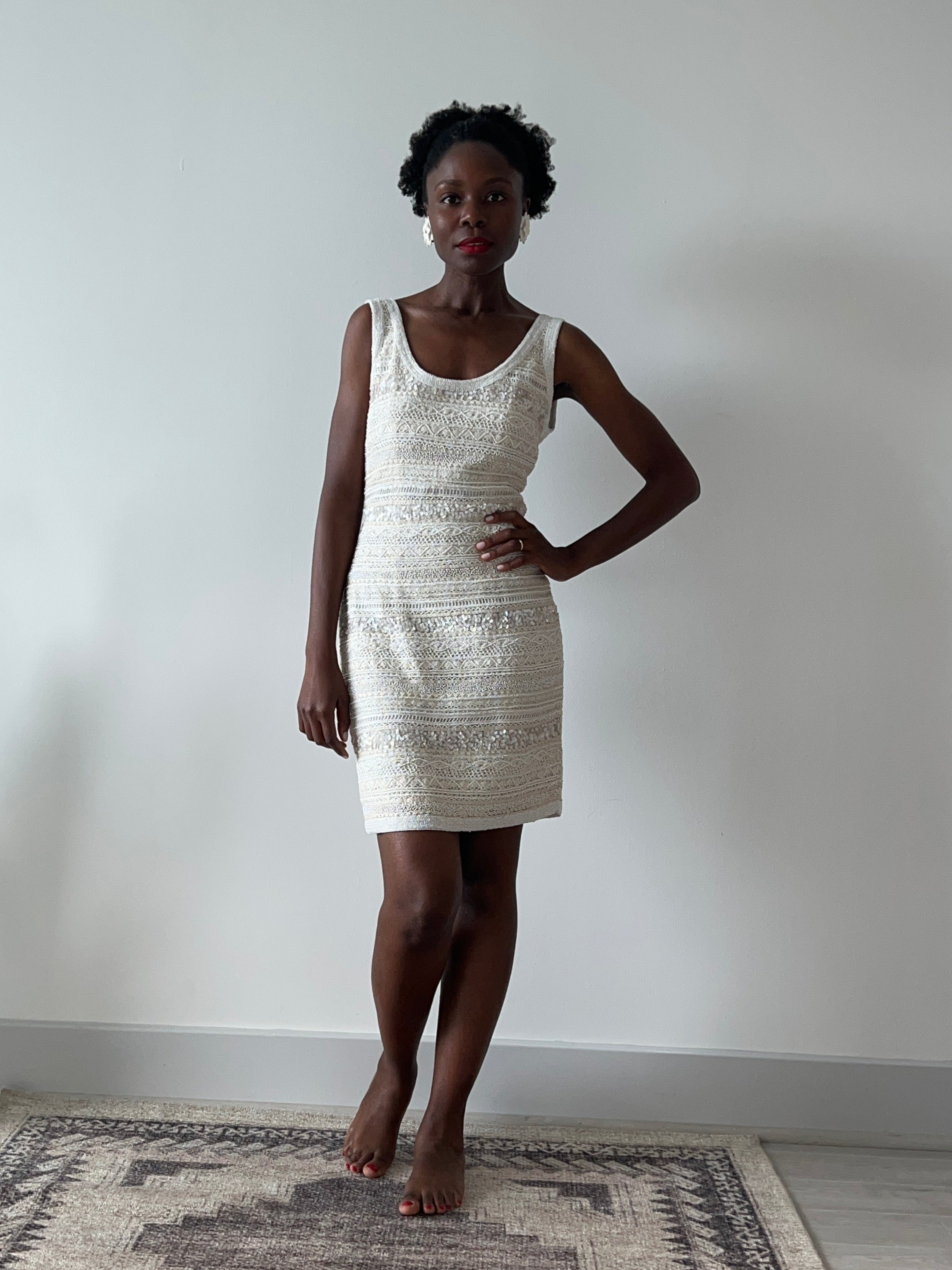 Person wearing a white dress standing against a plain wall with a patterned rug on the right.