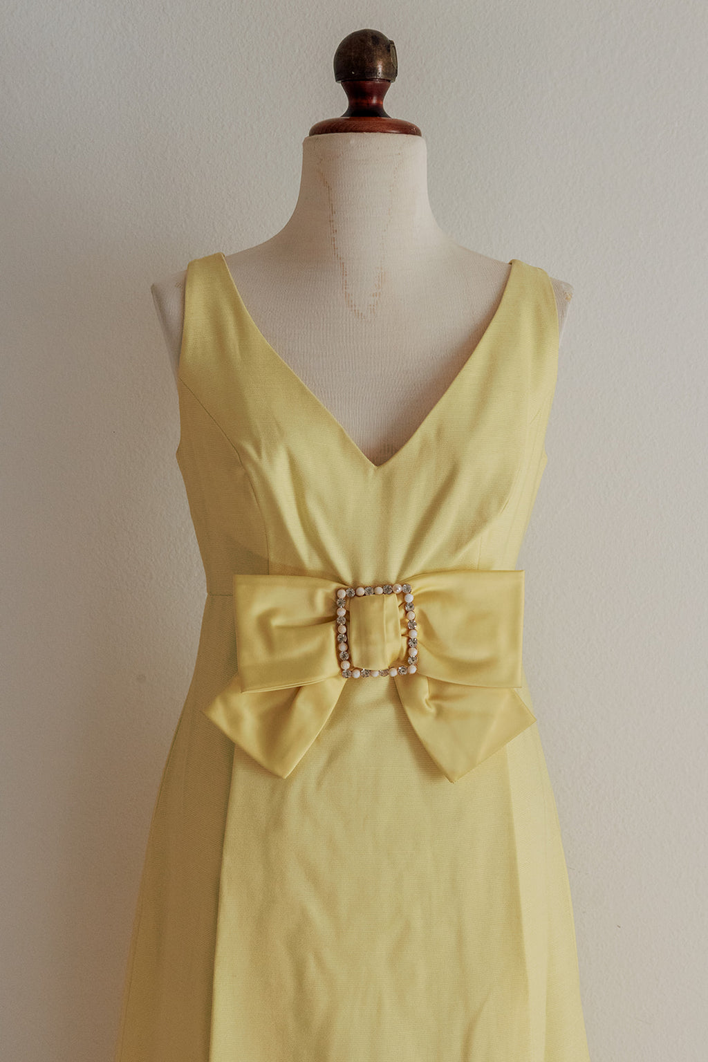 Yellow dress with a large bow and decorative buckle on a mannequin against a plain background